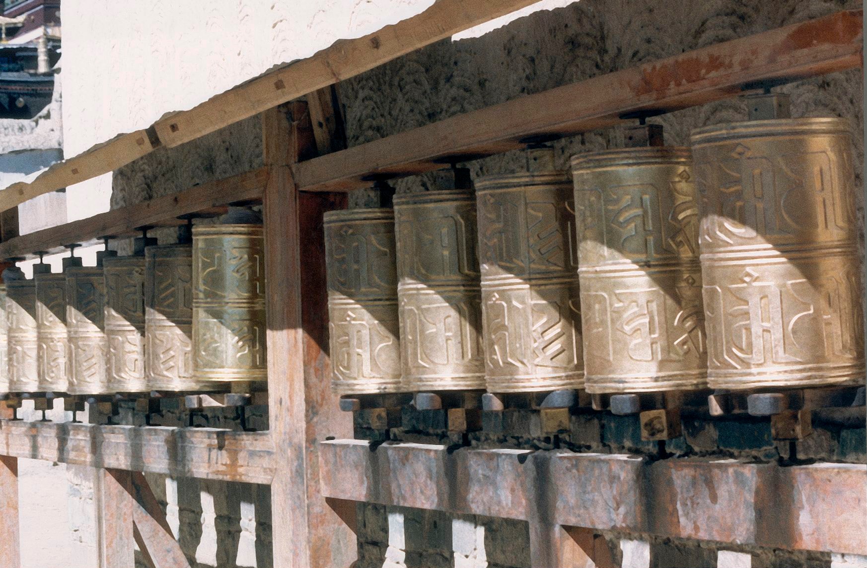Rows of brass prayer wheels mounted on a wooden structure, sunlight casting long shadows across the inscribed cylinders